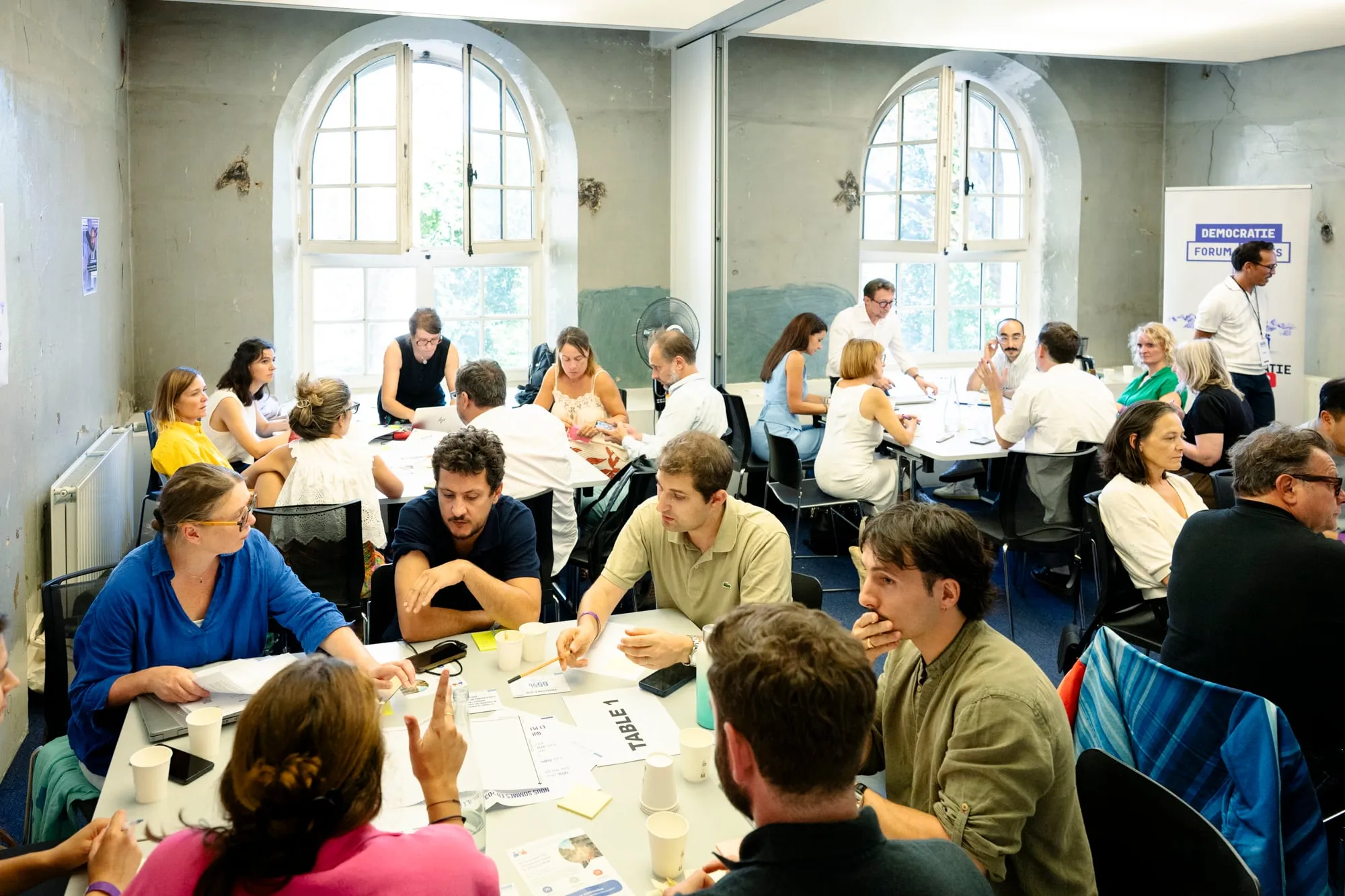 Participants en atelier autour de tables de travail, dans une salle aux fenêtres cintrées.