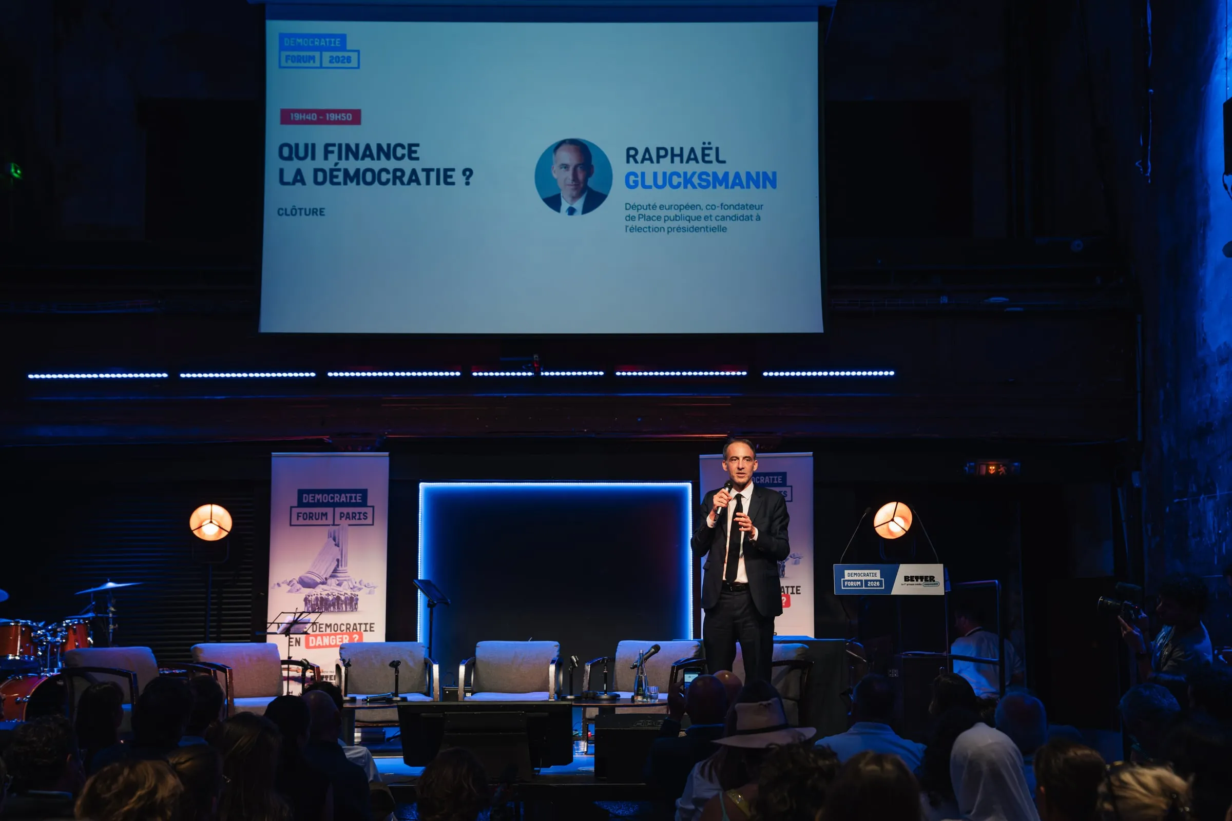 Raphaël Glucksmann au micro sur la scène, devant l'écran « Qui finance la démocratie ? ».