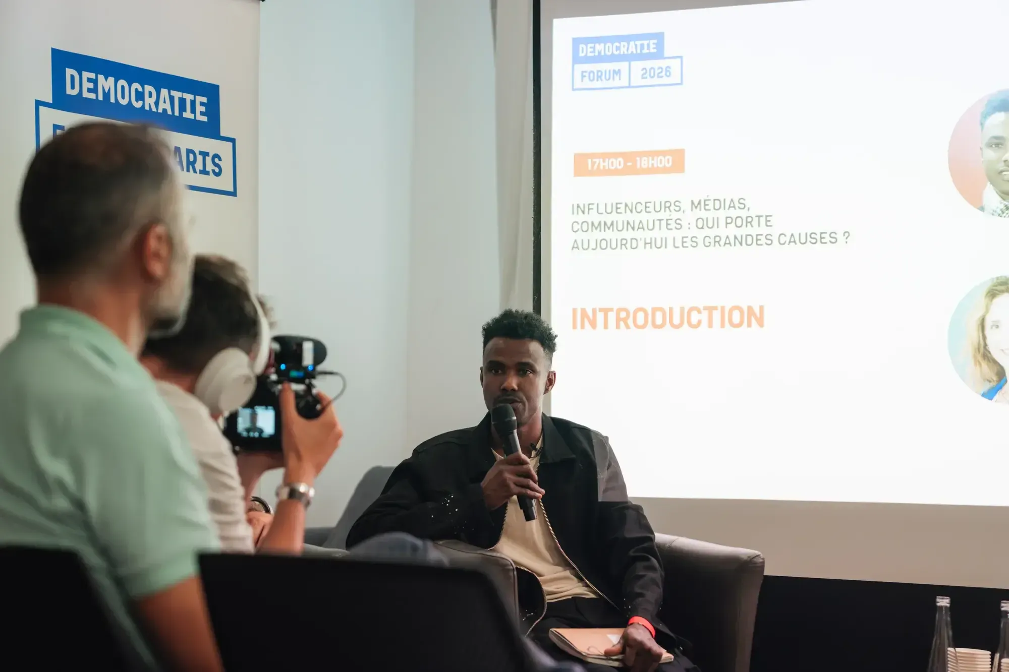 Un intervenant au micro dans un fauteuil, devant l'écran de la table ronde « Influenceurs, médias, communautés ». Un caméraman le filme au premier plan.