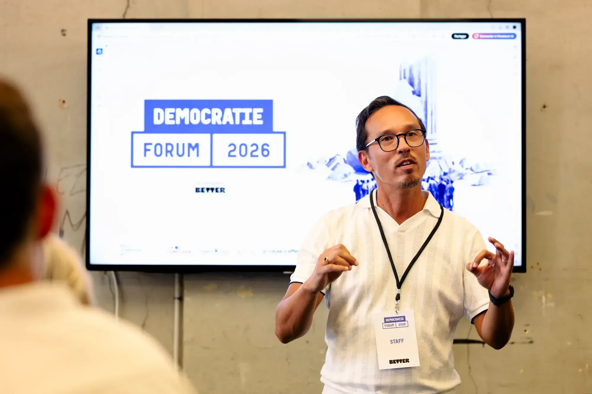 Un membre de l'équipe, badge « staff » au cou, présente la page d'accueil du Démocratie Forum affichée sur un grand écran.