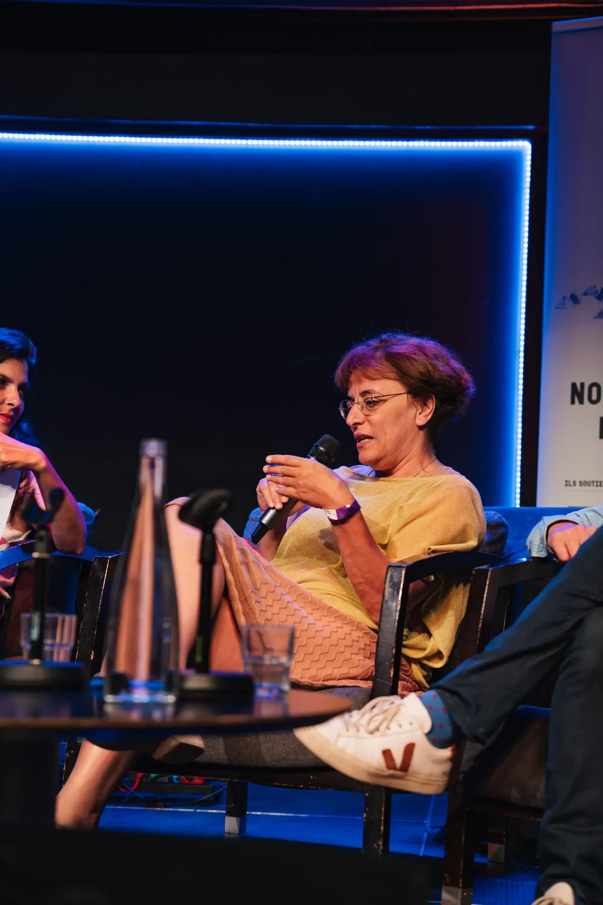 Une intervenante au micro sur la scène du forum, dans la lumière bleue, une autre intervenante à côté d'elle.